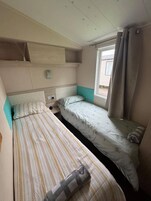 Una habitación pequeña y funcional con dos camas individuales, una ventana con cortinas y un estante en la pared.