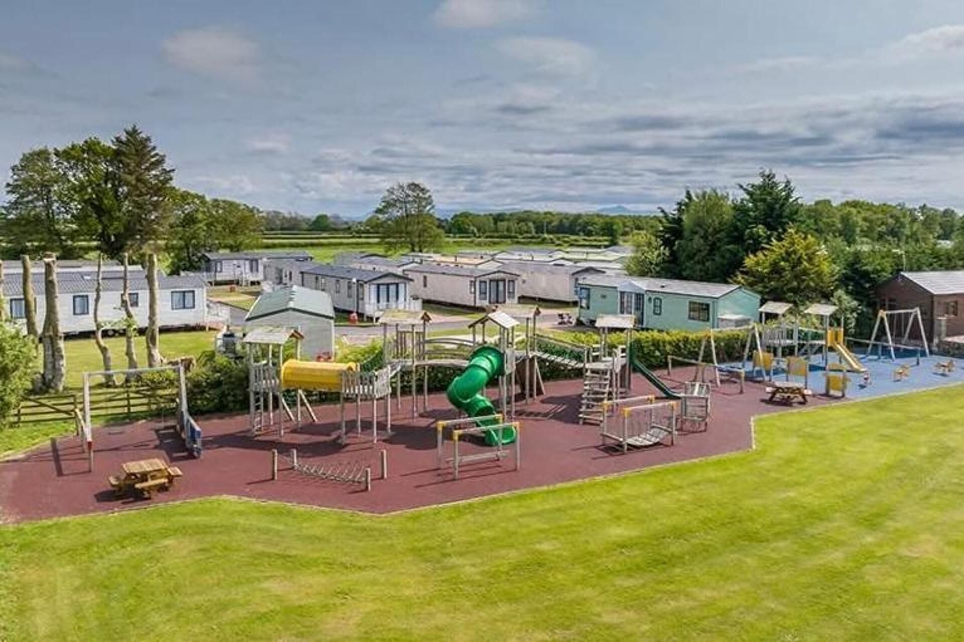 A playground with various play structures, including a green slide, surrounded by a grassy area and a row of mobile homes in the background.