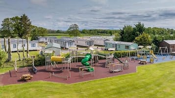 Un parque infantil con diversas estructuras de juego, incluyendo un tobogán verde, rodeado de césped y una fila de viviendas móviles al fondo.