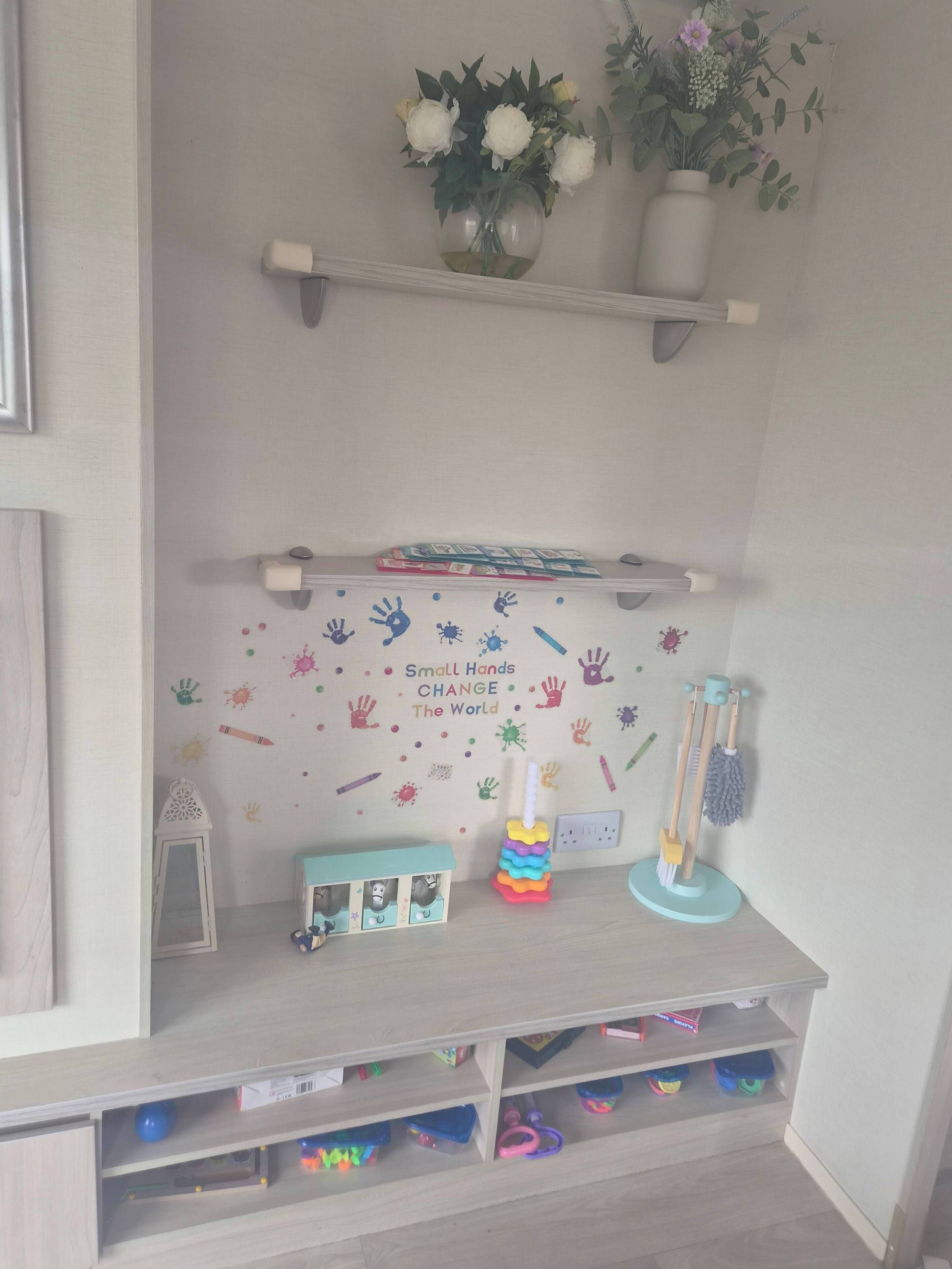 Habitación infantil con un mural de huellas de manos y una cita motivadora, una estantería con juguetes y un pequeño escritorio.