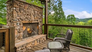 A stone fireplace with a fire, surrounded by wicker chairs and a table on a wooden deck with a view of a forested landscape. - CoveMtnComf: Pool, MtnViews, HotTub, Wifi, Pets, Theater, Games (Sevierville)