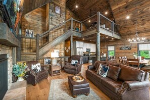 A spacious living area with a high ceiling, wooden beams, a staircase, and a kitchen in the background. - CoveMtnComf: Pool, MtnViews, HotTub, Wifi, Pets, Theater, Games (Sevierville)