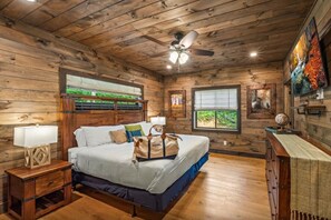 A wooden cabin bedroom with a bed, nightstands, a ceiling fan, and a window with a view of greenery. - CoveMtnComf: Pool, MtnViews, HotTub, Wifi, Pets, Theater, Games (Sevierville)