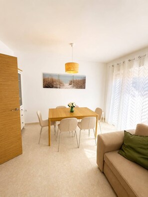 A modern living room with a wooden dining table, six chairs, a beige sofa with a green pillow, a hanging lamp, and a beach-themed artwork on the wall. - Townhouse located 50 meters from the beach in a family-friendly and very quiet neighborhood (Almassora)