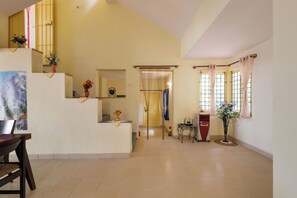 A spacious room with a high ceiling, a dining area with a table and chairs, a small kitchen area, and a large window with curtains. - Villa Terracotta by Redolivenr Art of Living3bhk (Bengaluru)