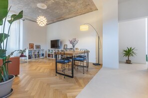 Ein modernes Wohnzimmer mit Holzboden, einer Essecke mit Tisch und Stühlen, einem Bücherregal, einem Fernseher und einer großen Pflanze.