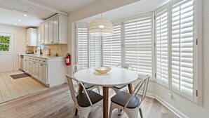 Eine moderne Küche mit weißen Schränken, ein Essbereich mit rundem Tisch und Stühlen sowie große Fenster mit weißen Fensterläden.