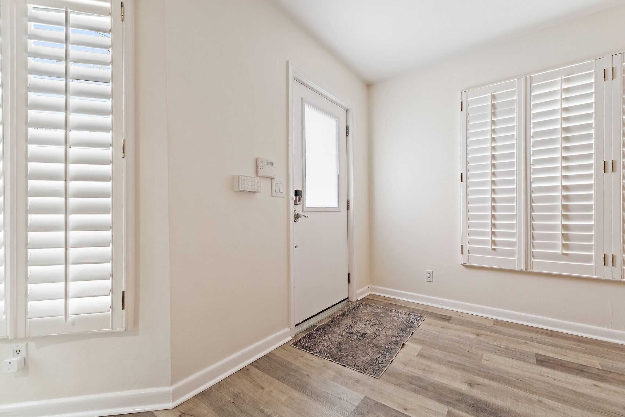A room with a door, a window with blinds, a wall-mounted thermostat, and a patterned rug on a wooden floor.