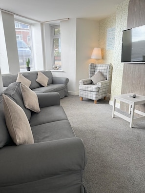 Ein modernes Wohnzimmer mit einem grauen Sofa, einem karierten Sessel, einem kleinen weißen Tisch und einem an der Wand befestigten Fernseher.