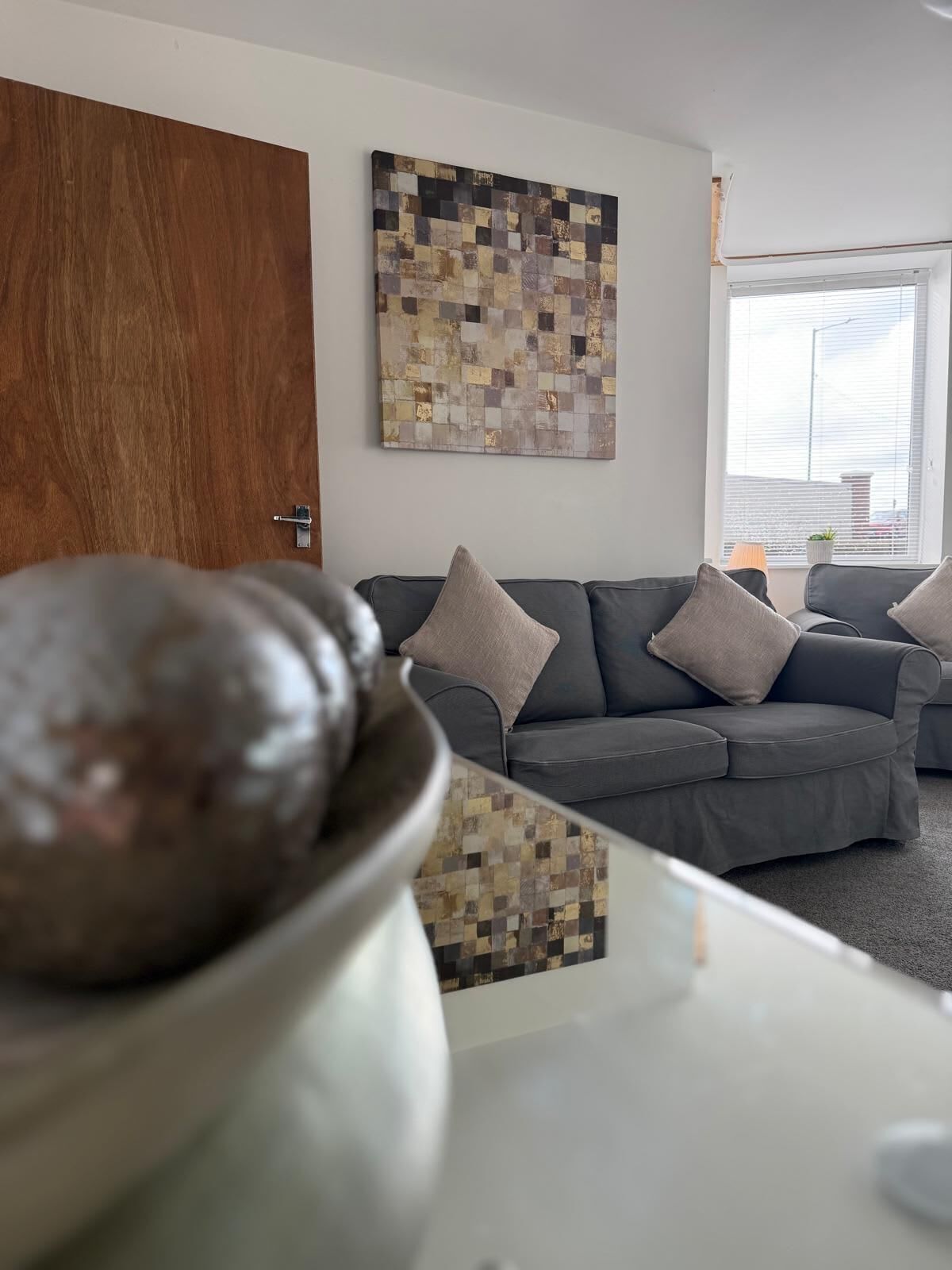 Un salon moderne avec un canapé gris, un élément décoratif mural et une fenêtre équipée de stores.