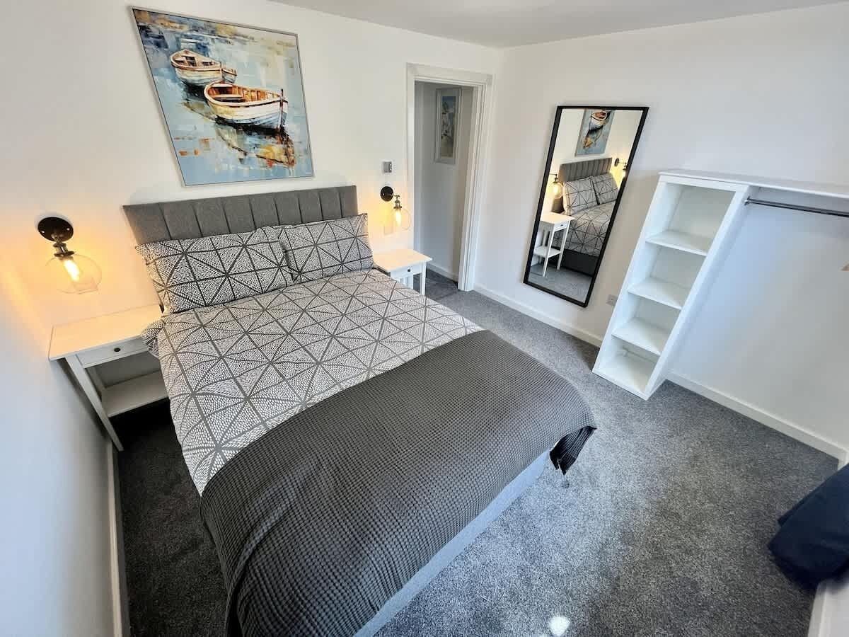 A bedroom with a bed, bedside tables, a nightstand, a mirror, and a wall-mounted shelf.
