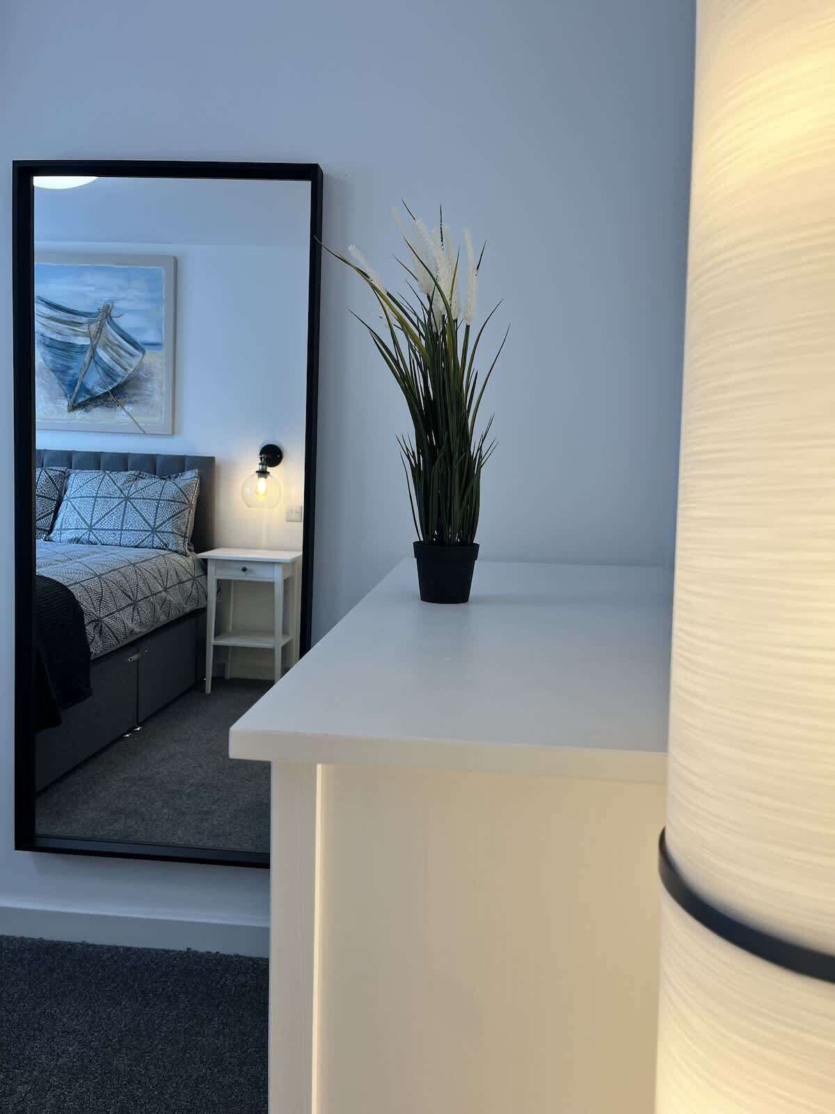 A bedroom with a bed, a bedside table, a lamp, a mirror, and a potted plant.