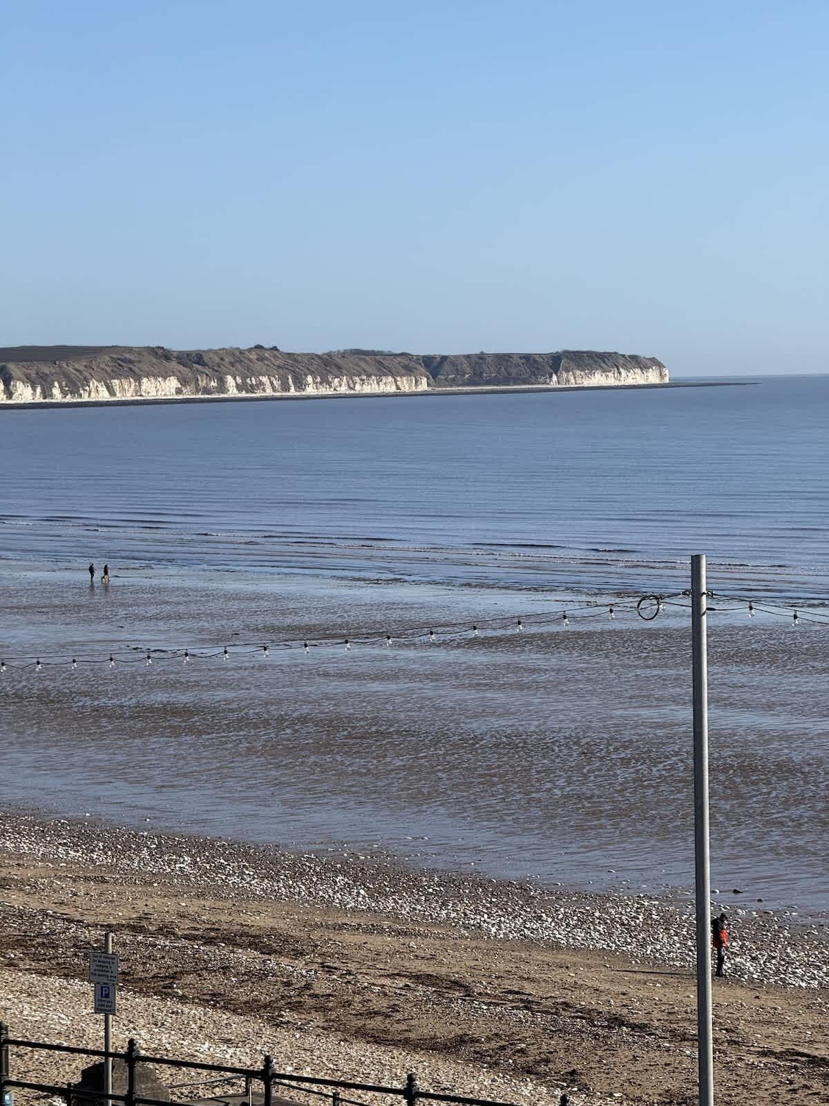 A coastal view with a rocky beach, a clear sky, and a distant cliff formation.