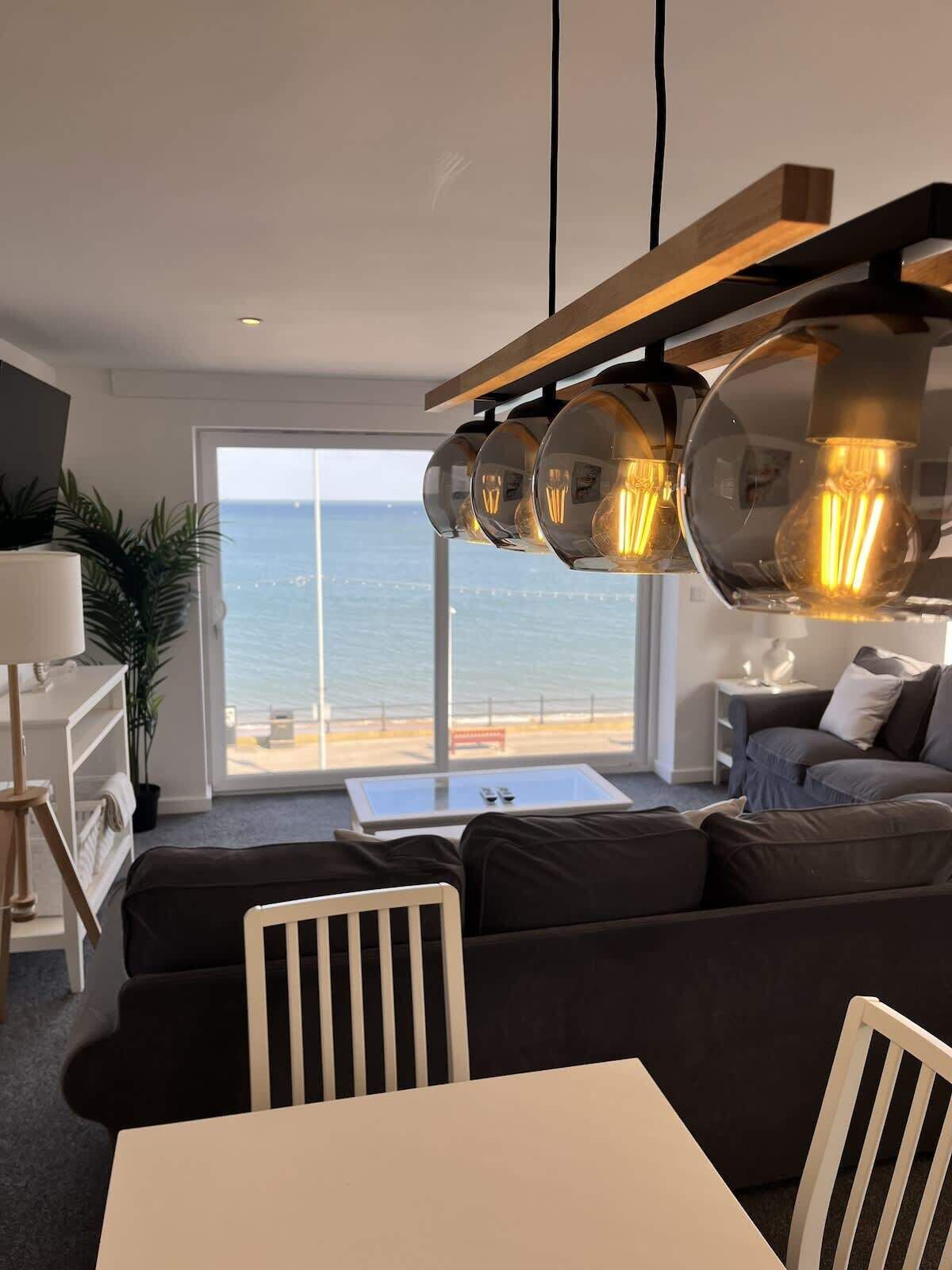 A modern living room with a large window overlooking the sea, a sofa set, a dining table, and a unique pendant light fixture.