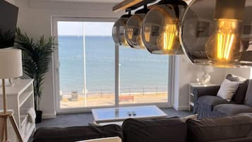 A modern living room with a large window overlooking the sea, a sofa set, a dining table, and a unique pendant light fixture.