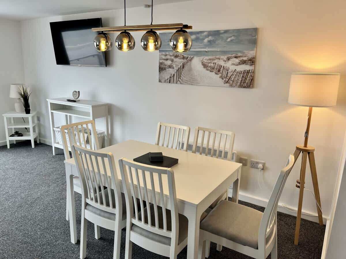 A dining area with a white table and chairs, a mounted flat-screen TV, a painting on the wall, and a floor lamp.