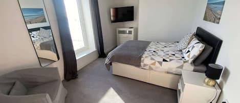 Un dormitorio con cama, mesita de noche, una silla, televisor y ventana con cortinas.