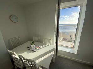Une pièce avec une table, des chaises, une horloge et une vue sur la plage par la fenêtre.