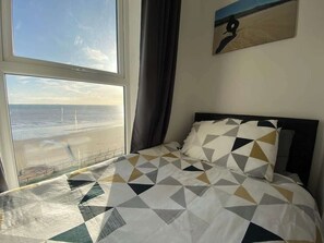Una cama con una colcha de estampado geométrico, vista al mar a través de una ventana y un cuadro en la pared.