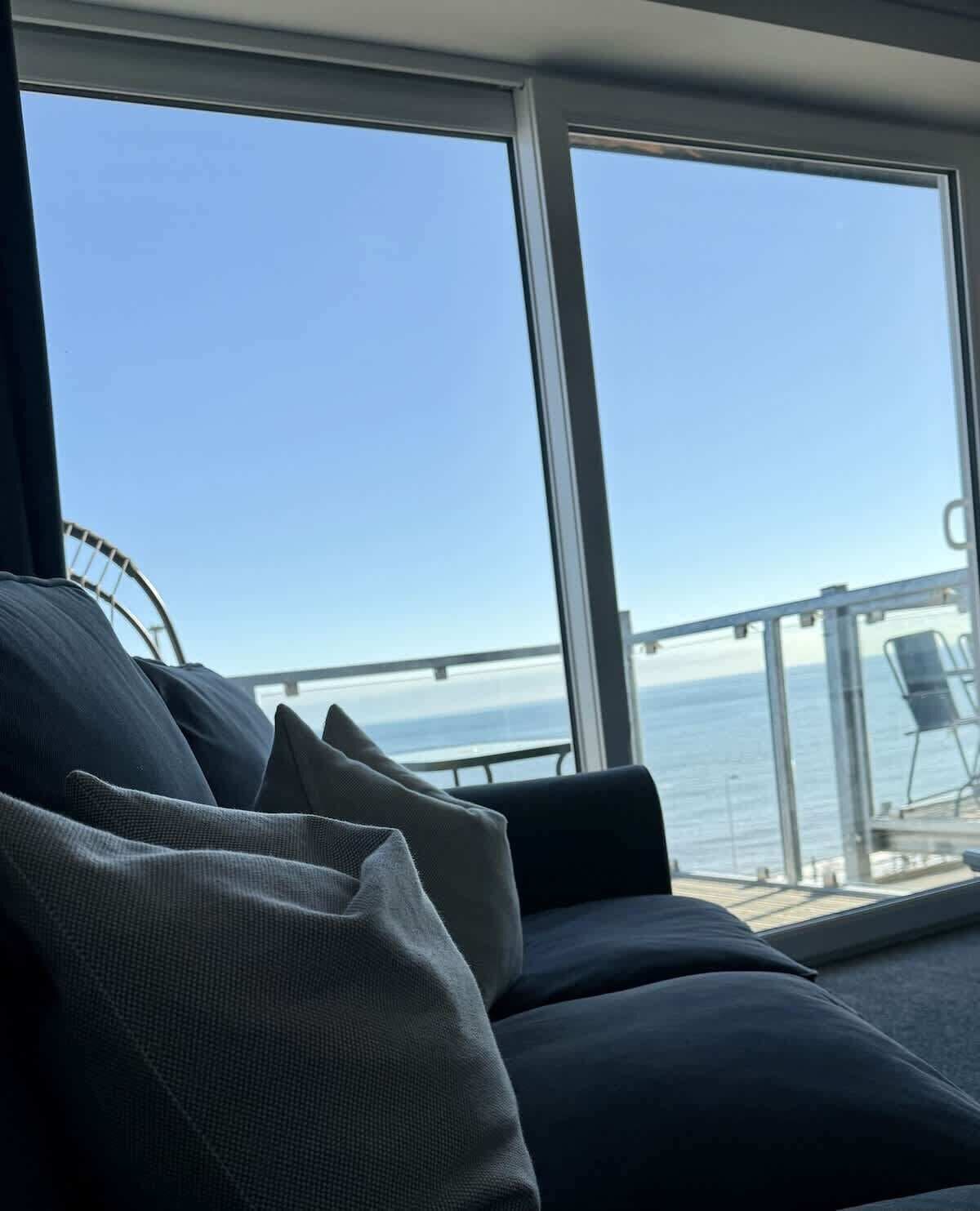 A room with a large window overlooking the sea, a sofa with cushions, and a glass railing.