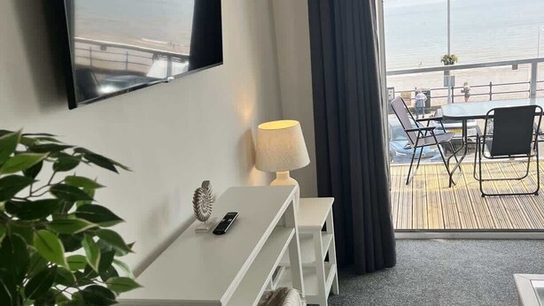 Quarto de hotel com uma cama, escrivaninha, cadeira, TV fixada na parede e vista para o mar pela janela.