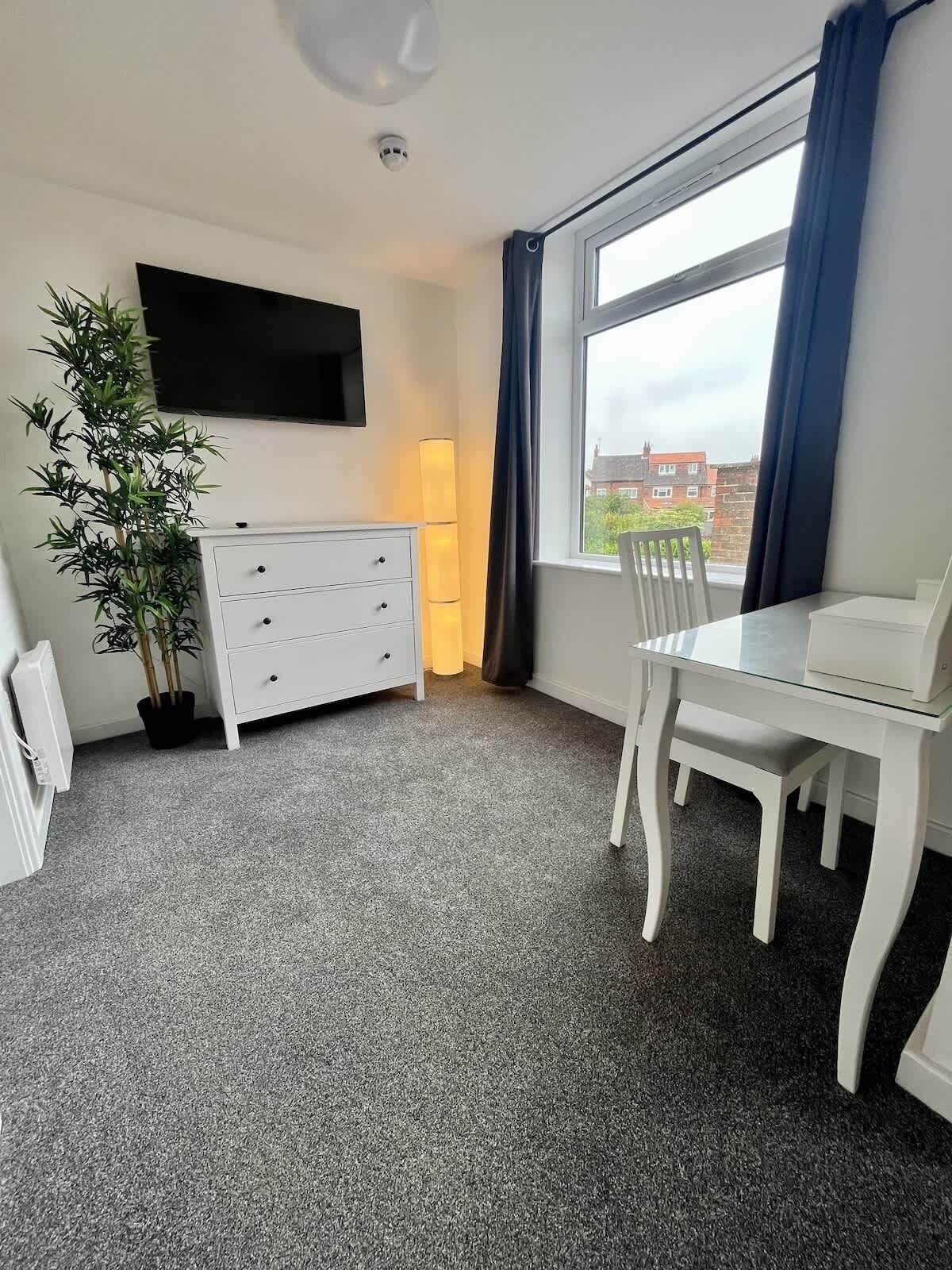 Una habitación con un aparador blanco, un televisor montado en la pared, una mesa de comedor con sillas y un amplio ventanal con cortinas.