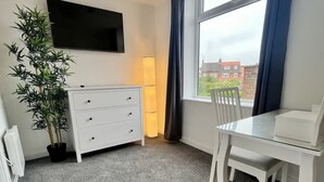 Um quarto com cômoda branca, televisão presa na parede, mesa de jantar com cadeiras e uma janela grande com cortinas.