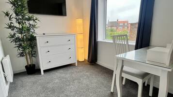 Una habitación con un aparador blanco, un televisor montado en la pared, una mesa de comedor con sillas y un amplio ventanal con cortinas.