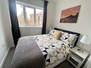 Ein Schlafzimmer mit einem Bett, einem Nachttisch, einer Lampe, einem Fenster und einem Bild an der Wand.