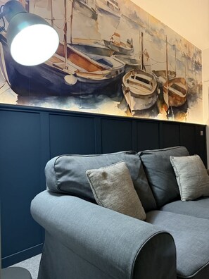 Ein Zimmer mit einer dunkelblauen Wand, einem grauen Sofa mit Kissen und einem großen Gemälde von Booten.