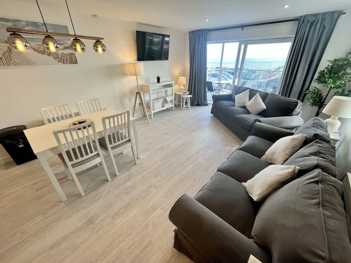 A modern living room with a sofa, a dining table with chairs, a TV mounted on the wall, and a large window with a view.