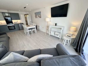 Un salon moderne avec un canapé gris, un coin repas avec des chaises blanches, une cuisine avec des meubles gris et un téléviseur à écran plat fixé au mur.