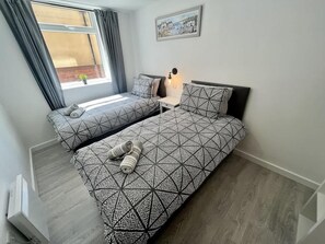 Une chambre avec deux lits, une table de chevet, une lampe, une fenêtre avec des rideaux et un tableau au mur.