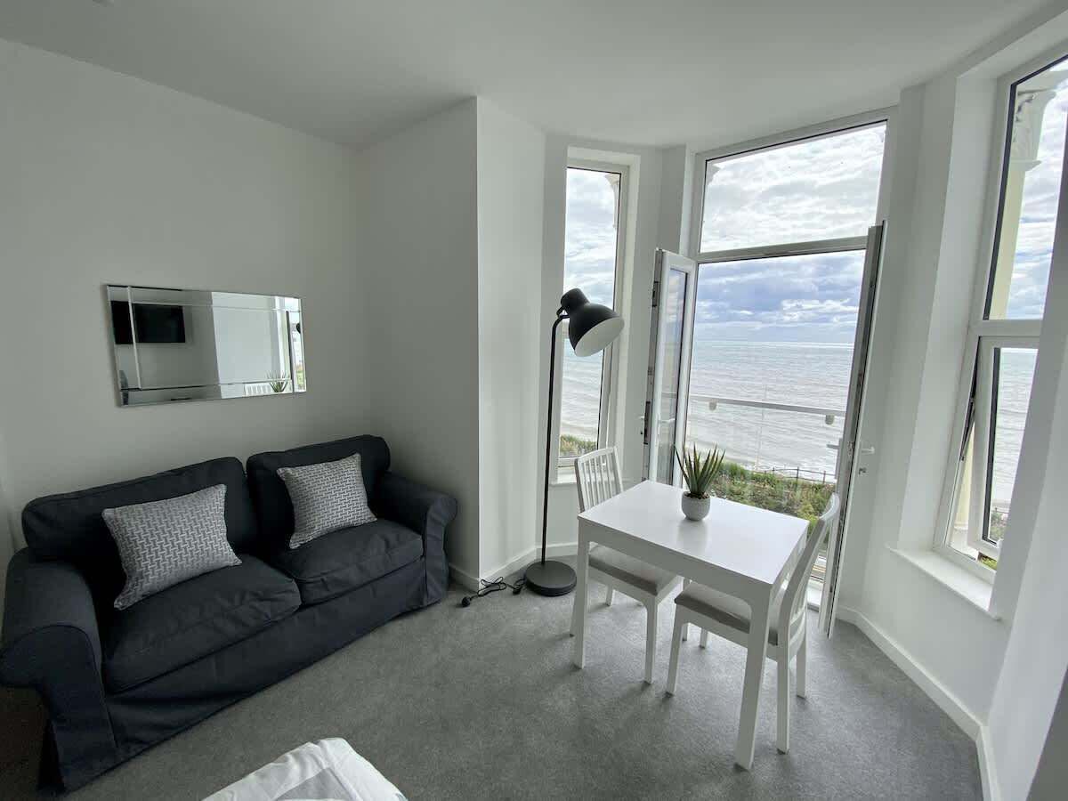 A modern living room with a grey sofa, a white dining table, and a view of the sea through large windows.