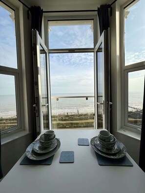A room with a table set for two, overlooking a coastal view with a fence and greenery. - Bridlington Bay Apt. 5 (East Riding of Yorkshire)