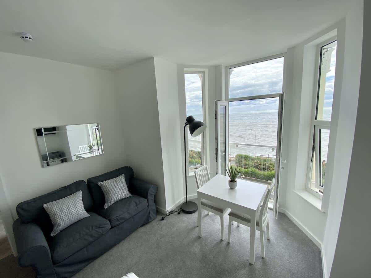 A room with a white table, a grey sofa, and a view of the sea through large windows.