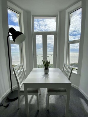 A room with a white table, two white chairs, a floor lamp, and a potted plant. There are three windows with views of the sea. - Bridlington Bay Apt. 5 (East Riding of Yorkshire)