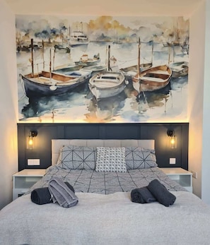 A bedroom with a large painting of boats on the wall, a patterned bedspread, and two bedside lamps. - Bridlington Bay Apt. 5 (East Riding of Yorkshire)