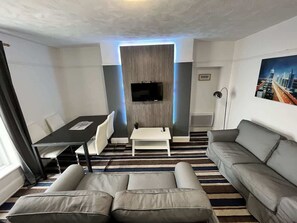 A modern living room with a flat-screen TV mounted on a wooden panel, a grey sofa, a striped rug, and a dining area with a table and chairs. - Harbour View Apt, Oceana (East Riding of Yorkshire)