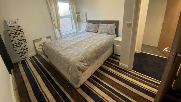 A bedroom with a bed, a bedside table, a nightstand, a window with curtains, and a striped carpet. - Harbour View Apt, Oceana (East Riding of Yorkshire)