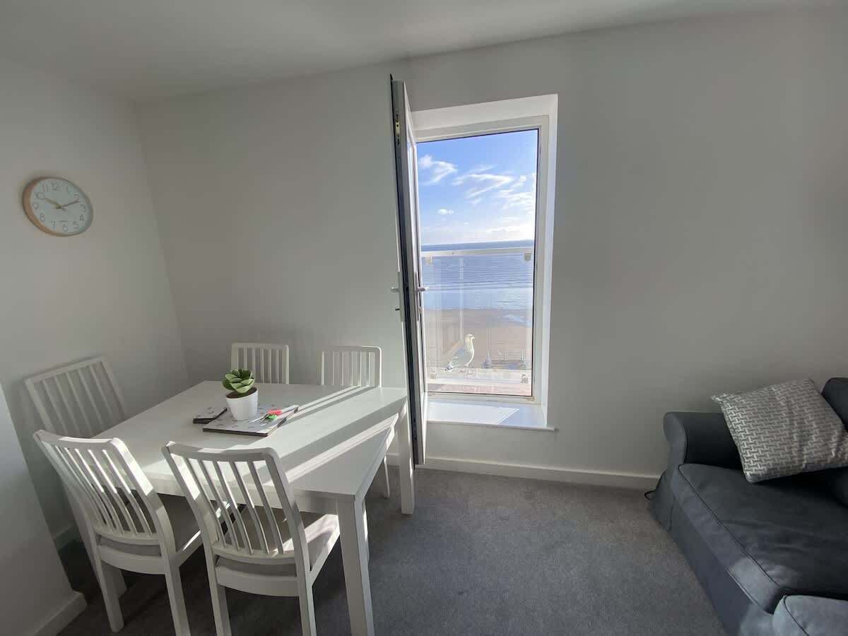 A room with a dining table, chairs, a small potted plant, a window with a view of the sea, and a grey sofa with a cushion.