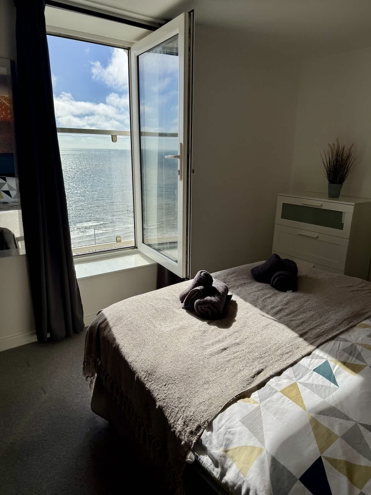 Habitación de hotel con cama, mesita de noche con planta y ventana con vista al mar.