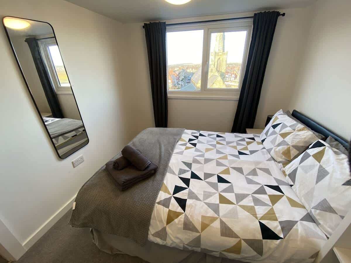 Ett hotellrum med en säng, ett fönster med gardiner, en spegel som hänger på väggen och utsikt över staden.