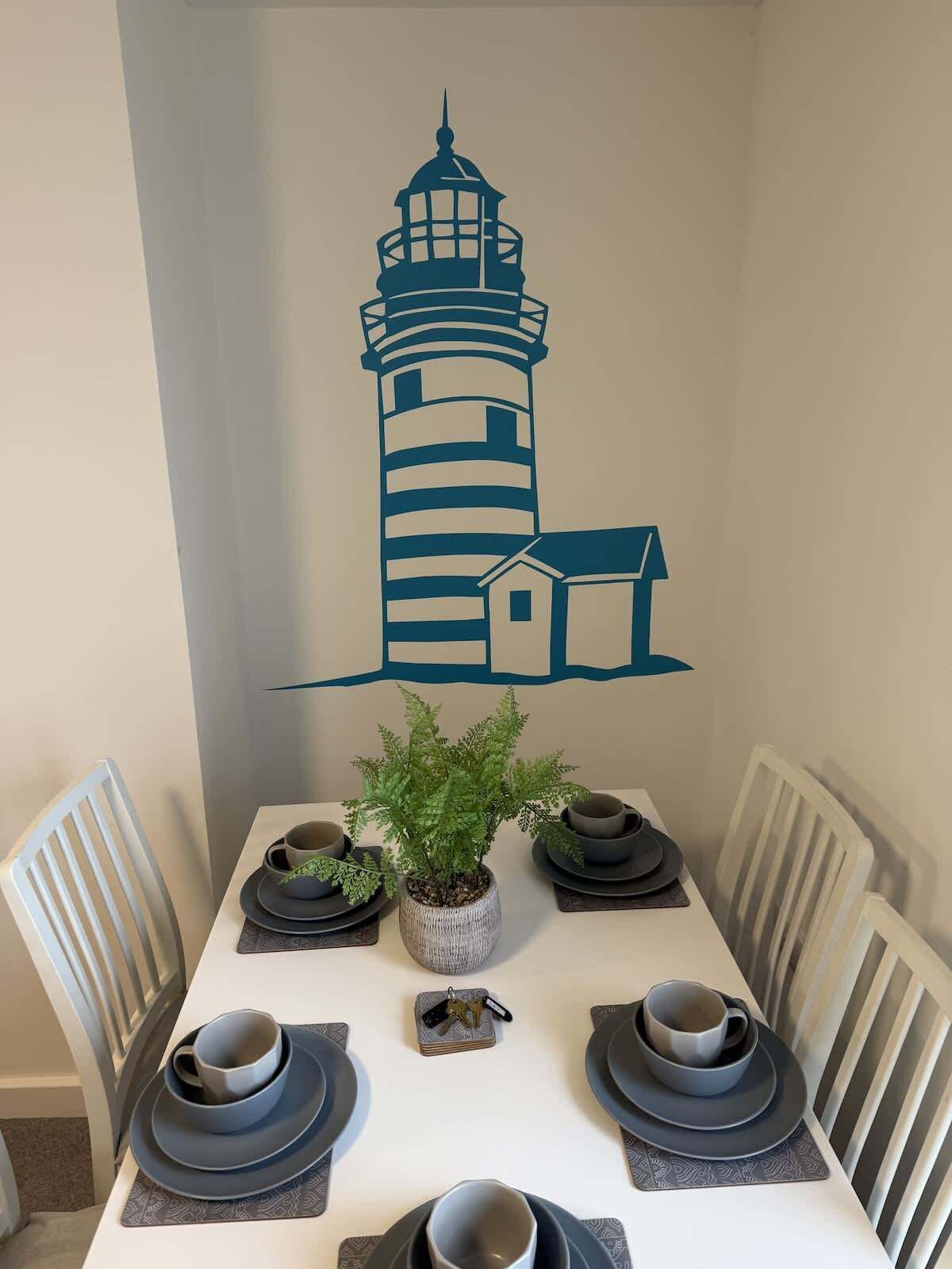 A dining table with a decorative wall mural of a lighthouse, a potted plant, and a set of black and white plates.