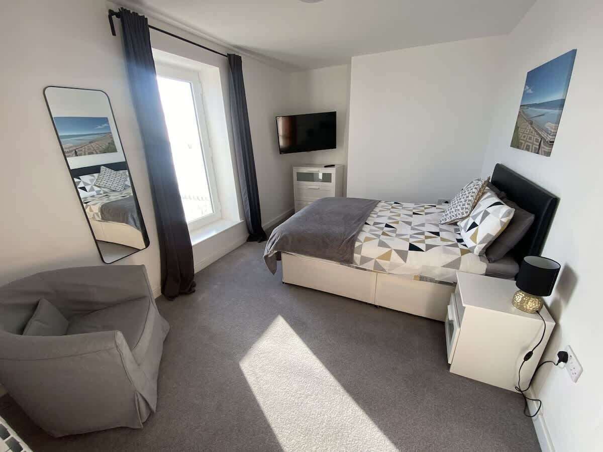 Ein Schlafzimmer mit Bett, Nachttisch, Fernseher, Stuhl und einem Fenster mit Vorhängen.