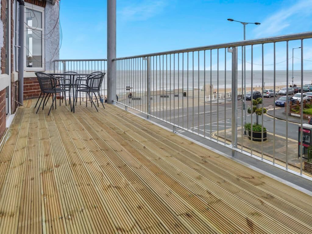 A balcony with a wooden floor, metal railing, and a view of the sea and parking area.
