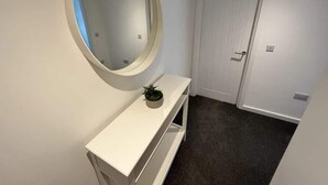 En hall med en vit dörr, ett vitt konsolbord med en spegel och en liten växt.