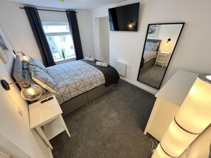A hotel room with a bed, bedside tables, a TV, a mirror, and a window with curtains. - Seaside Escapes Apt. 5 (East Riding of Yorkshire)