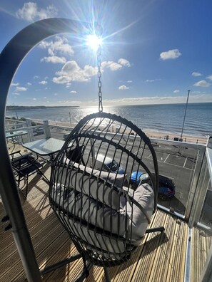 A sunlit deck with a hanging chair, a view of the sea, and a parking area. - Seaside Escapes Apt. 5 (East Riding of Yorkshire)