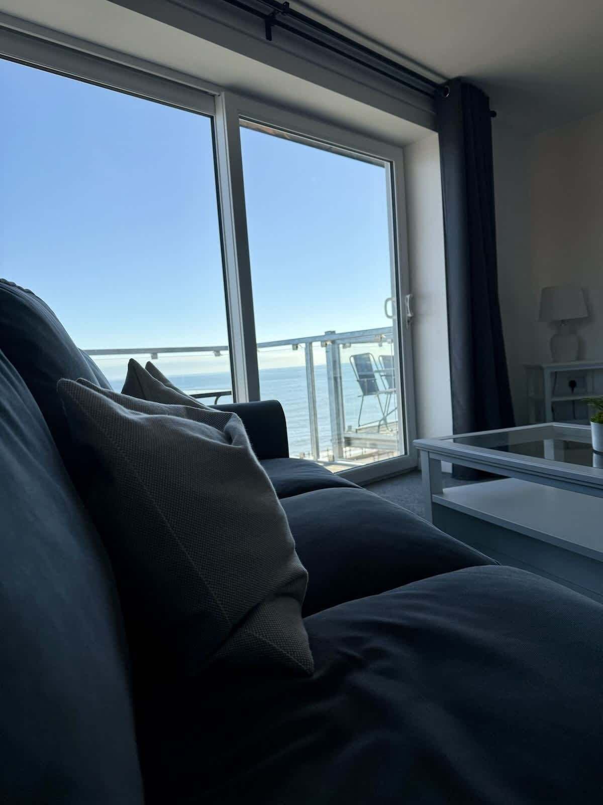 Ein modernes Wohnzimmer mit großem Fenster, einer Couch, einem Couchtisch und Blick auf das Meer und eine Brücke.